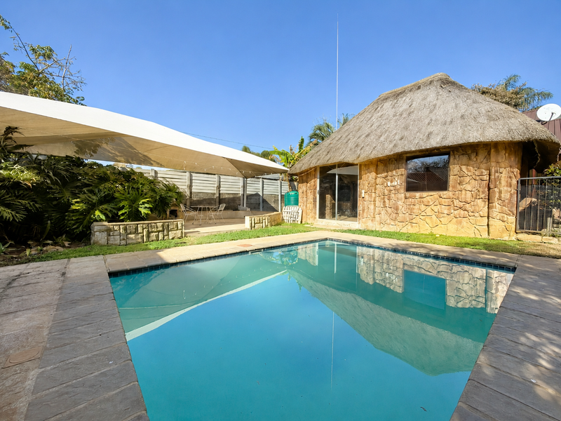 Outdoor pool area featuring a swimming pool, paved surround, shade sail, and a unique thatch-roofed structure with stone cladding.