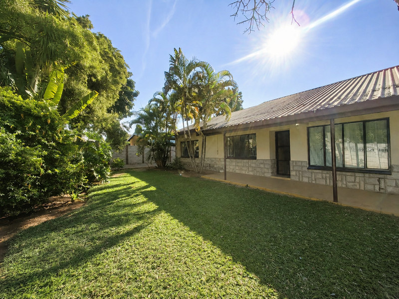 Rear view of the property featuring a covered patio, stone cladding, and a spacious garden area with established trees.