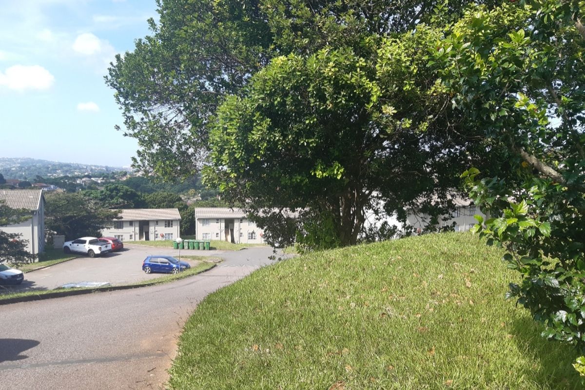 An exterior view of the apartment complex grounds featuring a paved driveway, landscaped lawn, and mature trees surrounding the residential buildings.