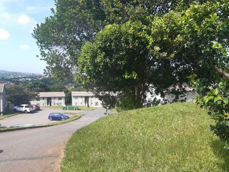 An exterior view of the apartment complex grounds featuring a paved driveway, landscaped lawn, and mature trees surrounding the residential buildings.