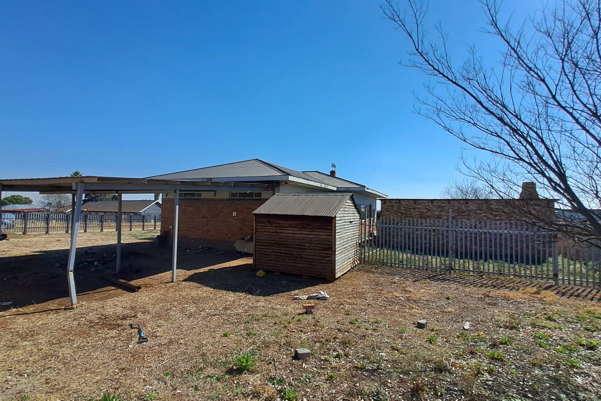 Rear view of the property featuring a brick house, a metal carport, a timber garden shed, and perimeter steel fencing on a dry yard.