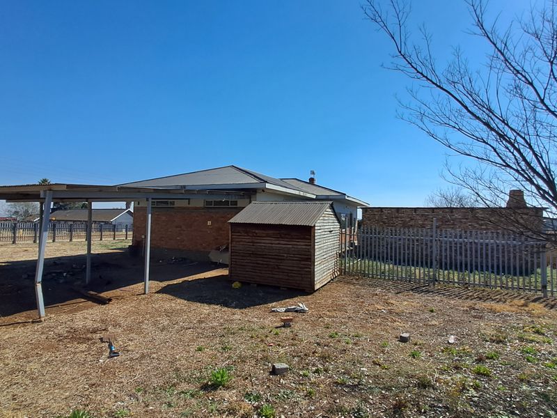 Rear view of the property featuring a brick house, a metal carport, a timber garden shed, and perimeter steel fencing on a dry yard.