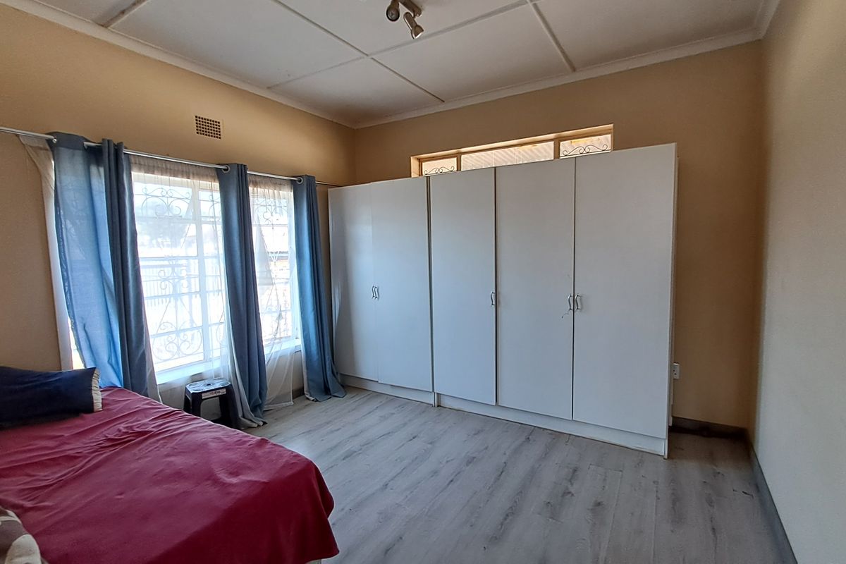 A bedroom featuring light-toned laminate flooring, built-in white cupboards, and security bars on the windows.