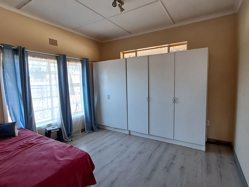 A bedroom featuring light-toned laminate flooring, built-in white cupboards, and security bars on the windows.