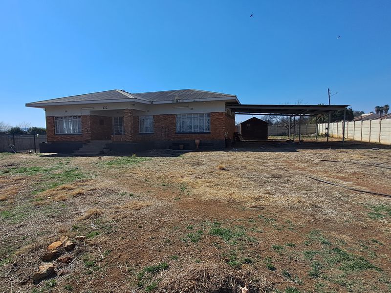 A single-story brick house featuring a metal roof, a side carport, and a large, undeveloped front yard enclosed by precast concrete boundary walls.