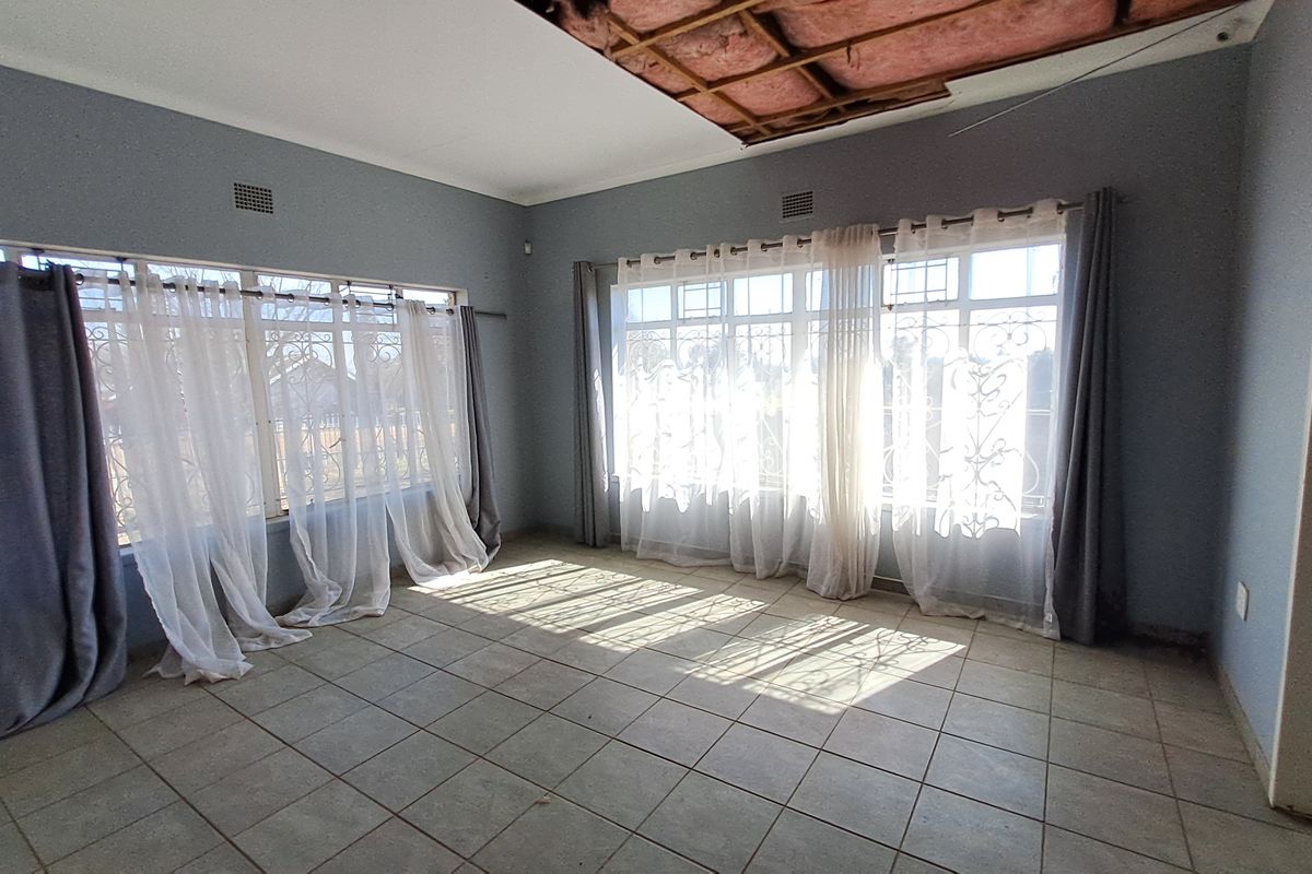 An empty living room featuring tiled floors and large windows with security bars, currently requiring ceiling repairs and maintenance.