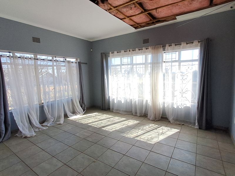 An empty living room featuring tiled floors and large windows with security bars, currently requiring ceiling repairs and maintenance.