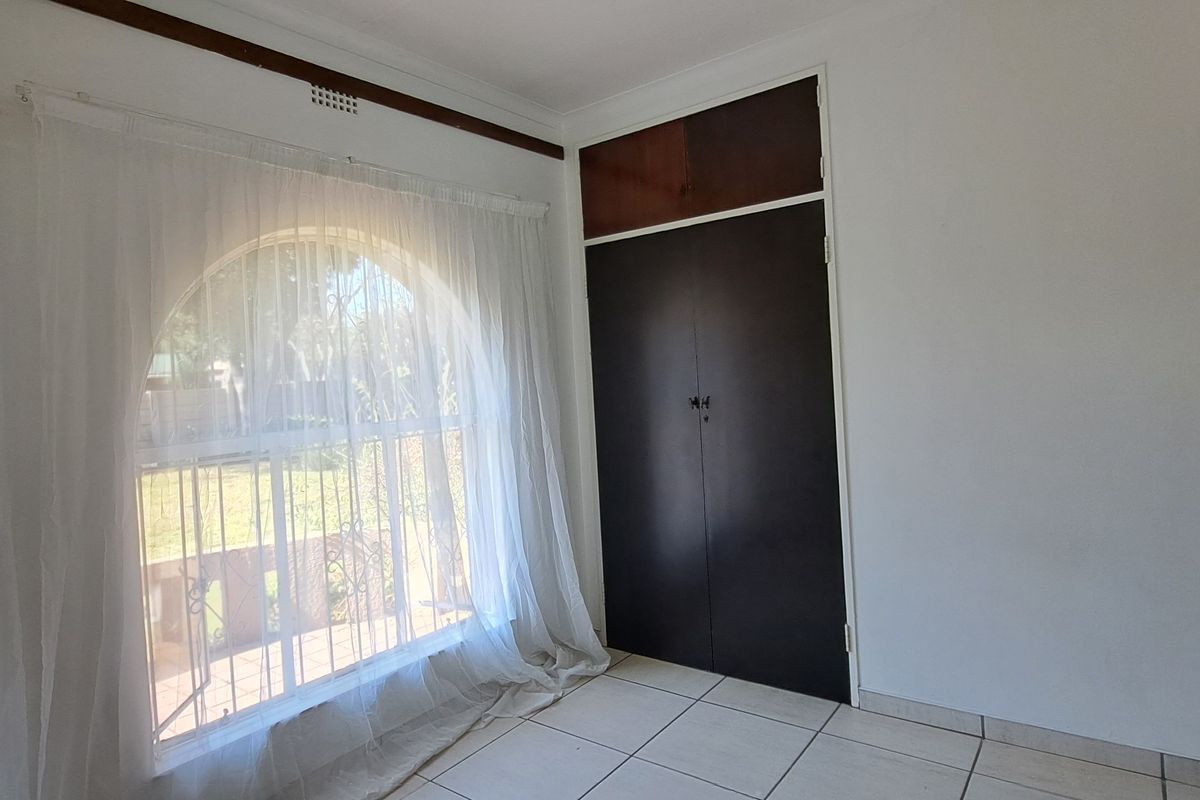 A bright, empty bedroom featuring tiled flooring, a large arched window with security bars, and a built-in cupboard with overhead storage.