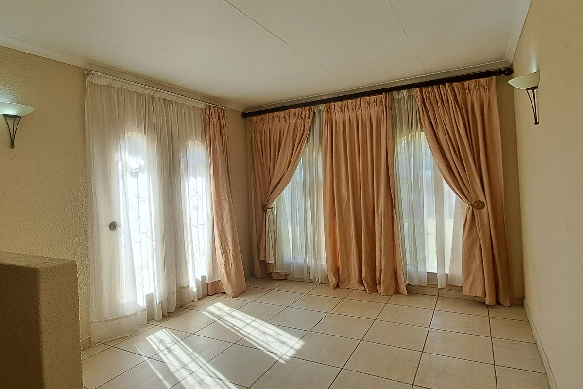 A bright, empty living room featuring tiled flooring, wall-mounted lighting, and large windows with existing curtain rods.