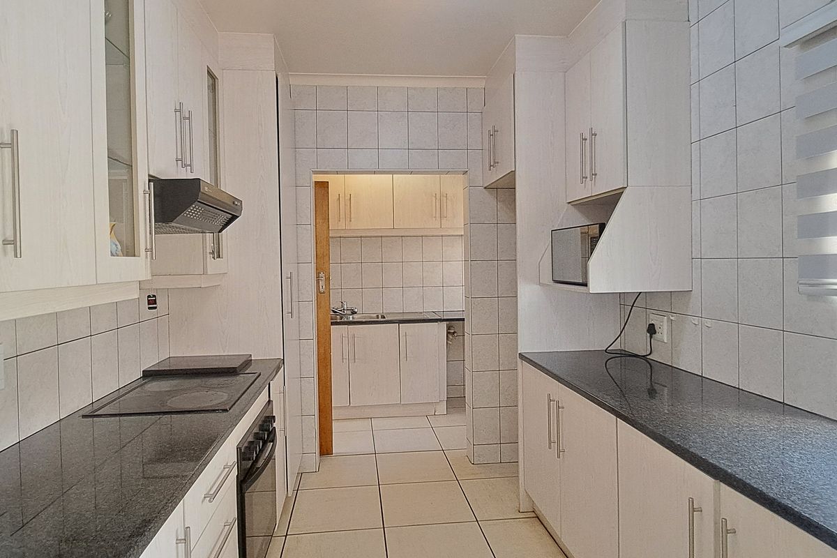 A clean, functional kitchen featuring white cabinetry, granite countertops, built-in appliances, and tiled finishes, with an adjoining scullery area visible.