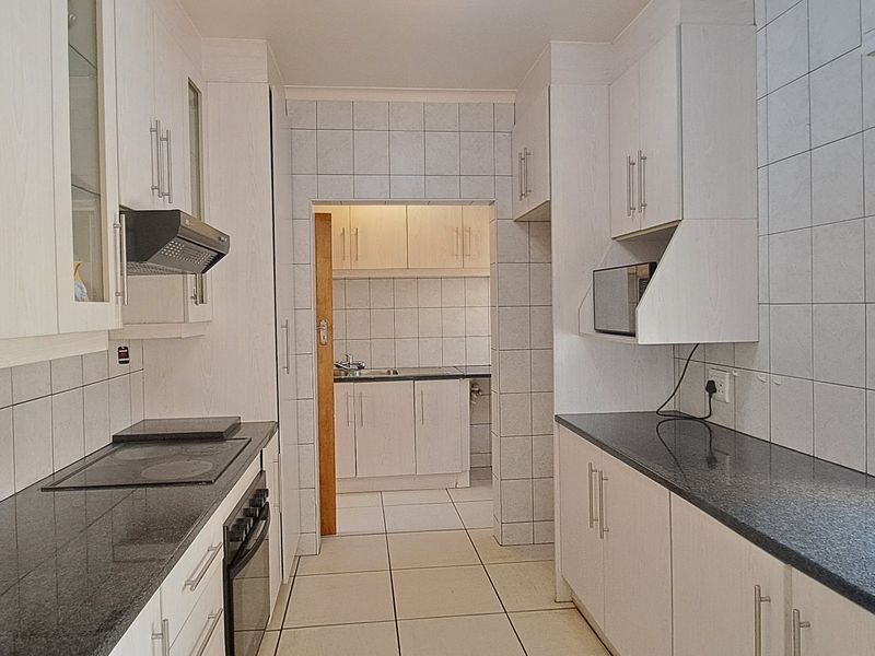 A clean, functional kitchen featuring white cabinetry, granite countertops, built-in appliances, and tiled finishes, with an adjoining scullery area visible.