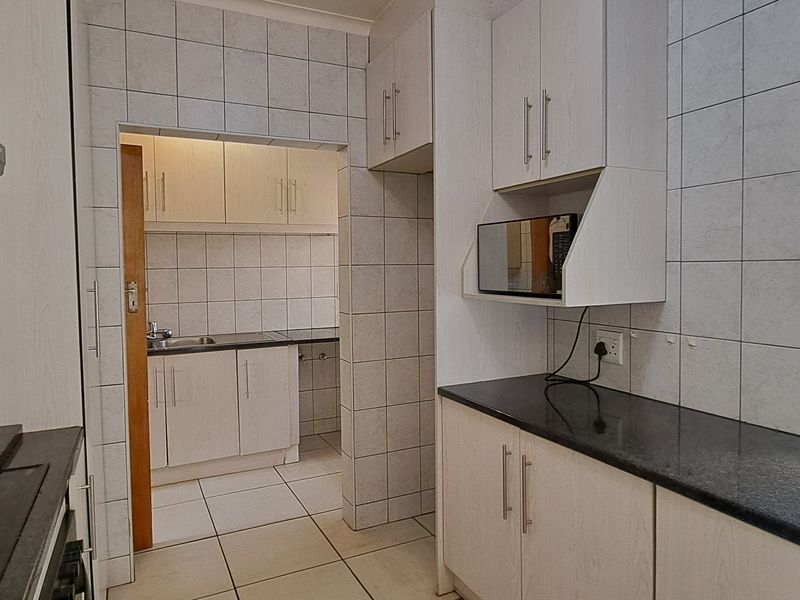 A functional kitchen featuring light wood cabinetry, dark granite countertops, tiled splashbacks, and an adjoining scullery area with additional storage and sink.