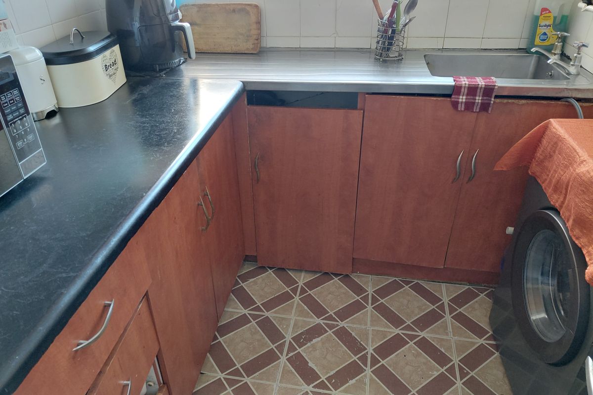 A functional kitchen featuring laminate cabinetry, a stainless steel sink, and tiled flooring, currently showing signs of wear and missing cabinet components.