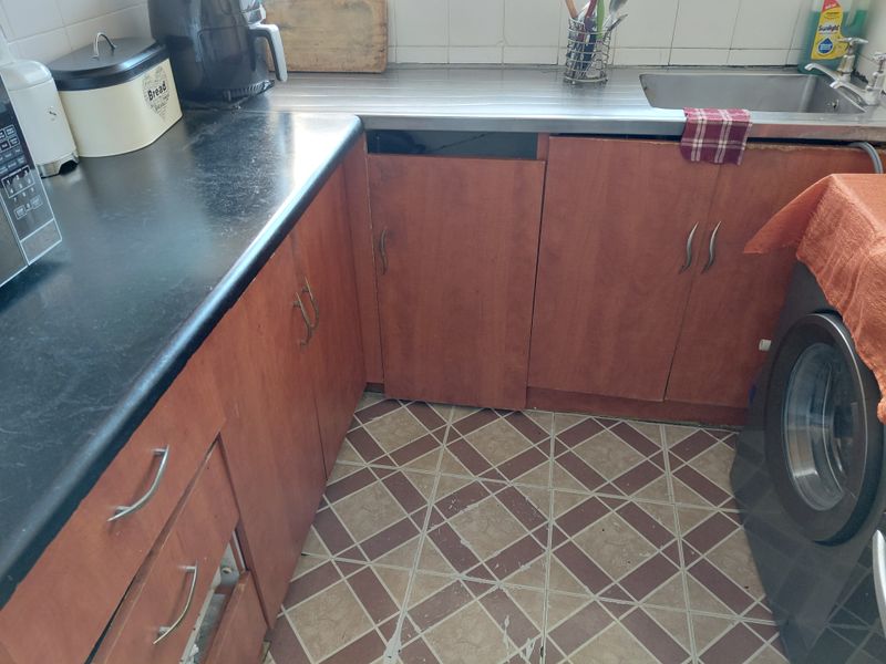 A dated kitchen featuring laminate cabinetry, a stainless steel sink, and tiled flooring, currently showing significant wear and missing cabinet components.