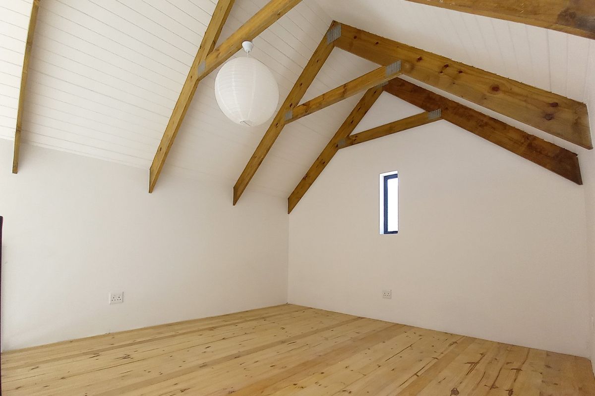 A bright, empty bedroom featuring exposed timber roof trusses, natural timber flooring, and a high vaulted ceiling with a vertical window.