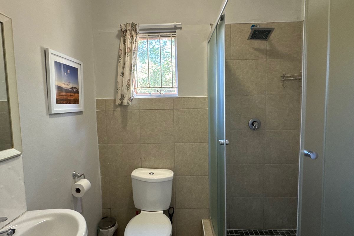 A functional bathroom featuring a shower enclosure, pedestal basin, and toilet with neutral wall tiling and security bars on the window.