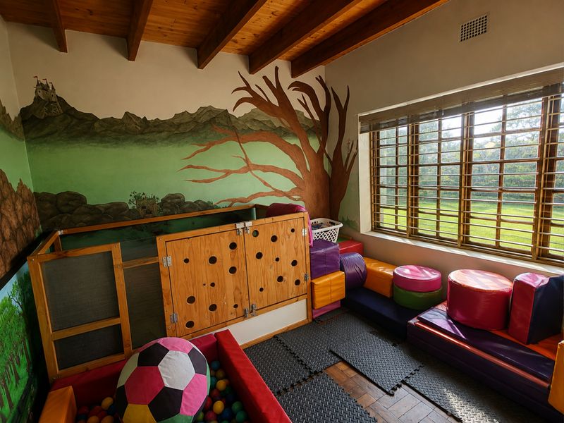 A bedroom featuring exposed timber ceiling beams, parquet flooring, and a large window with timber blinds, currently utilized as a children's play area.