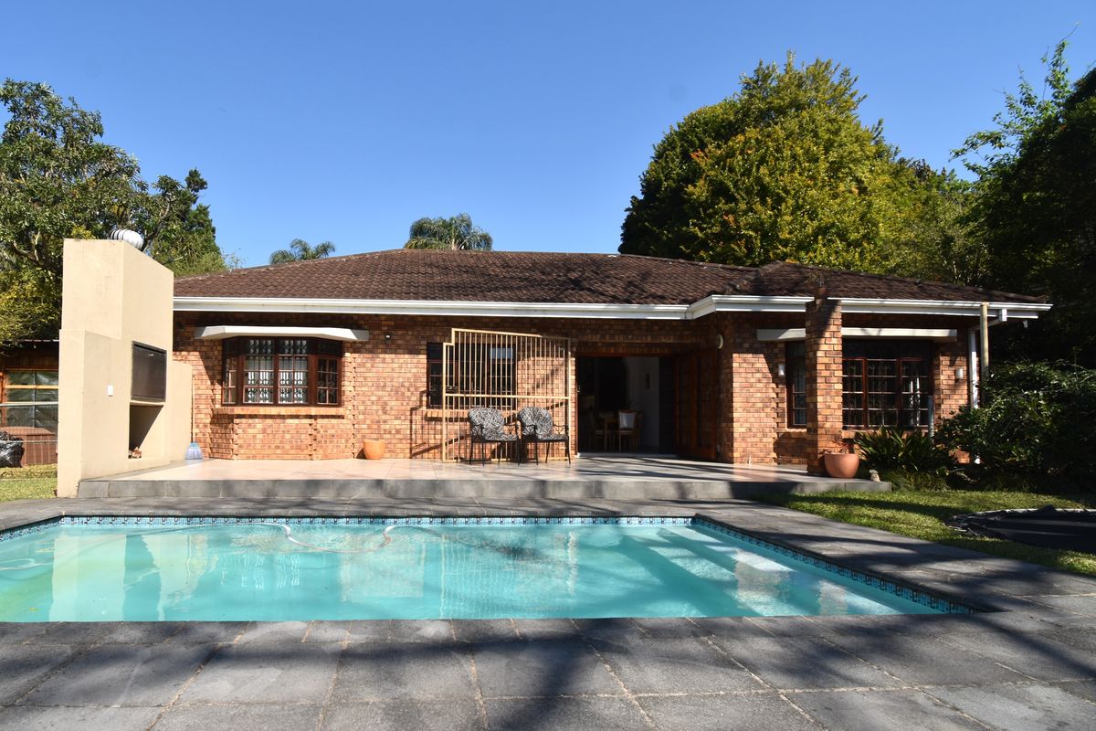 Rear view of a brick house featuring a swimming pool, paved patio area, and an outdoor fireplace, set within a landscaped garden.