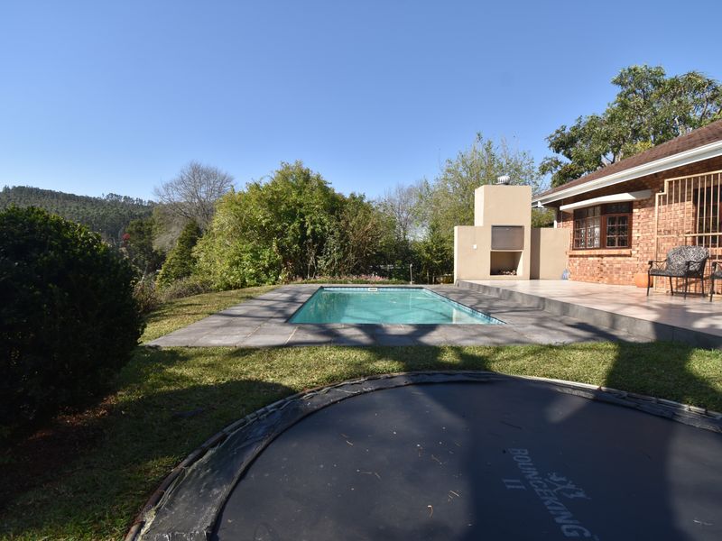 Outdoor pool area featuring a swimming pool, tiled patio, and a built-in braai, set against a brick house facade with garden surroundings.