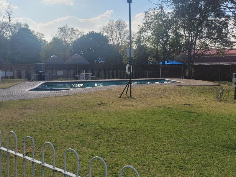 Communal pool area featuring a swimming pool, paved surround, and thatch umbrellas set within a fenced garden space.