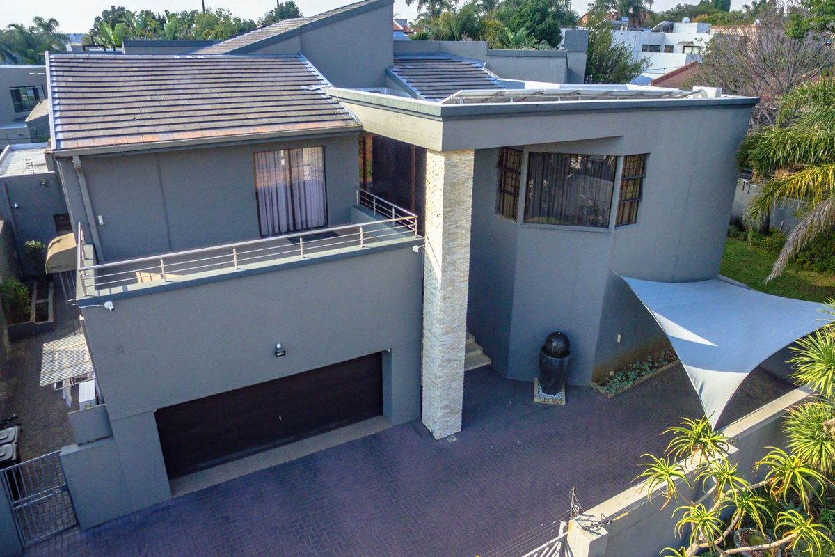 Aerial view of a modern double-story house featuring a paved driveway, double garage, balcony with metal railings, and a stone-clad architectural pillar.