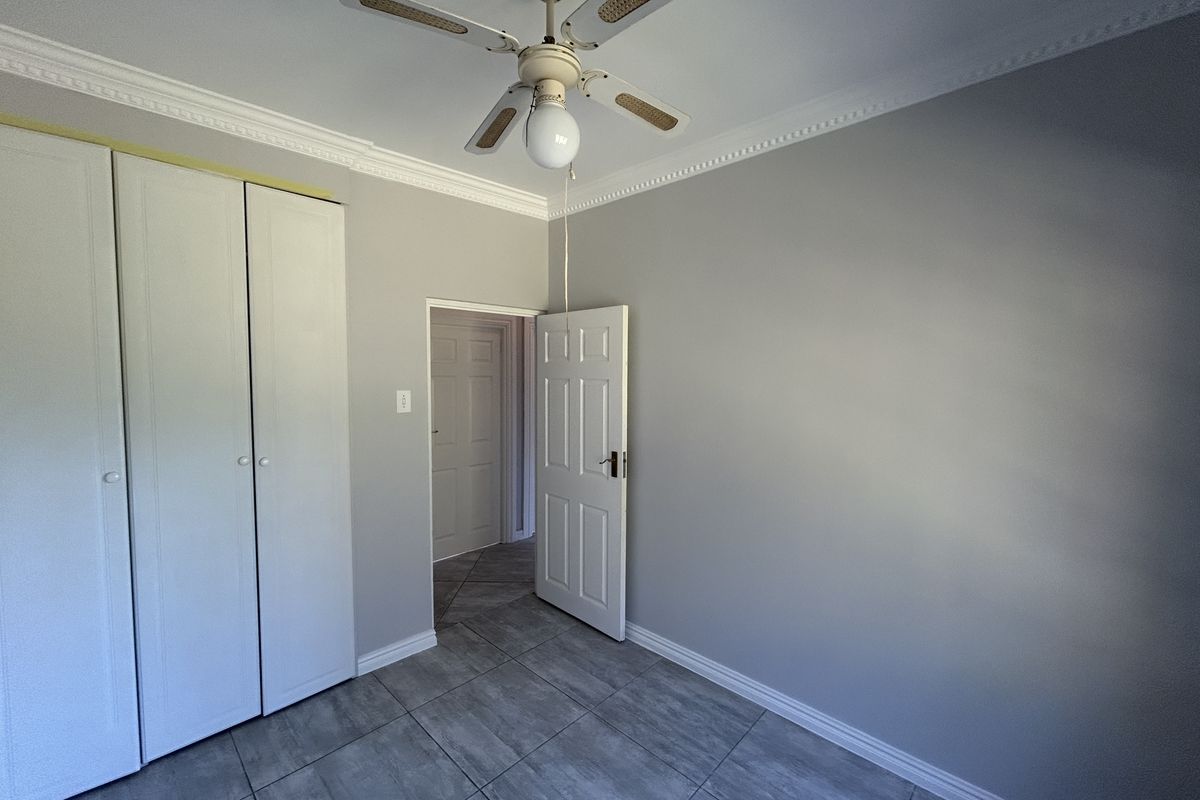 A clean, empty bedroom featuring neutral grey walls, tiled flooring, built-in cupboards, and a ceiling fan, offering a versatile space ready for furnishing.
