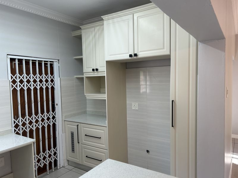 A clean, modern kitchen area featuring neutral built-in cabinetry, stone countertops, tiled backsplash, and a secure entrance gate.