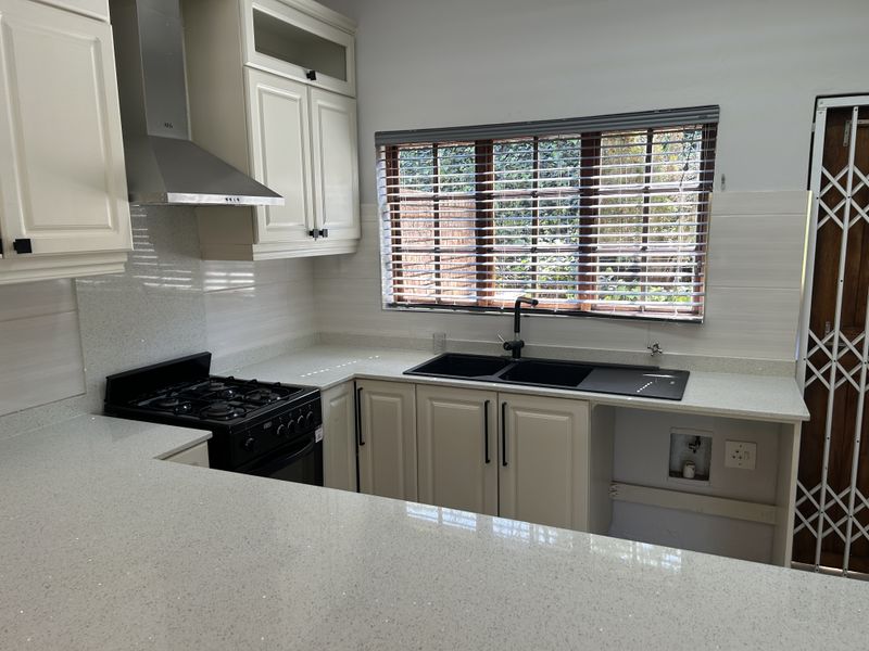 A clean, modern kitchen featuring light-toned cabinetry, quartz countertops, a gas stove, extractor fan, and a double sink with a view of the garden.