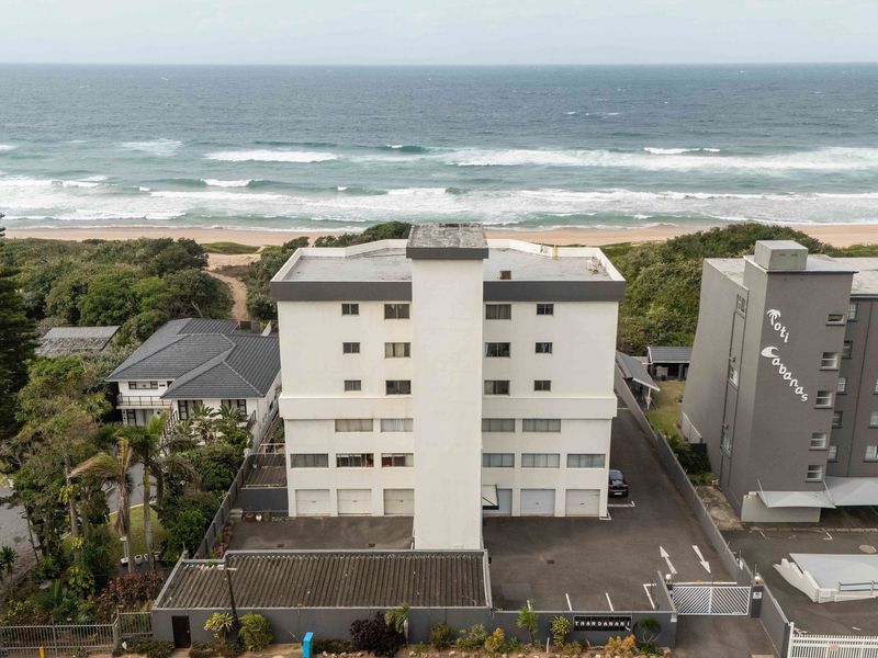 Aerial view of a multi-story apartment building situated directly on the beachfront, featuring a paved driveway, secure gated access, and ground-level garages.