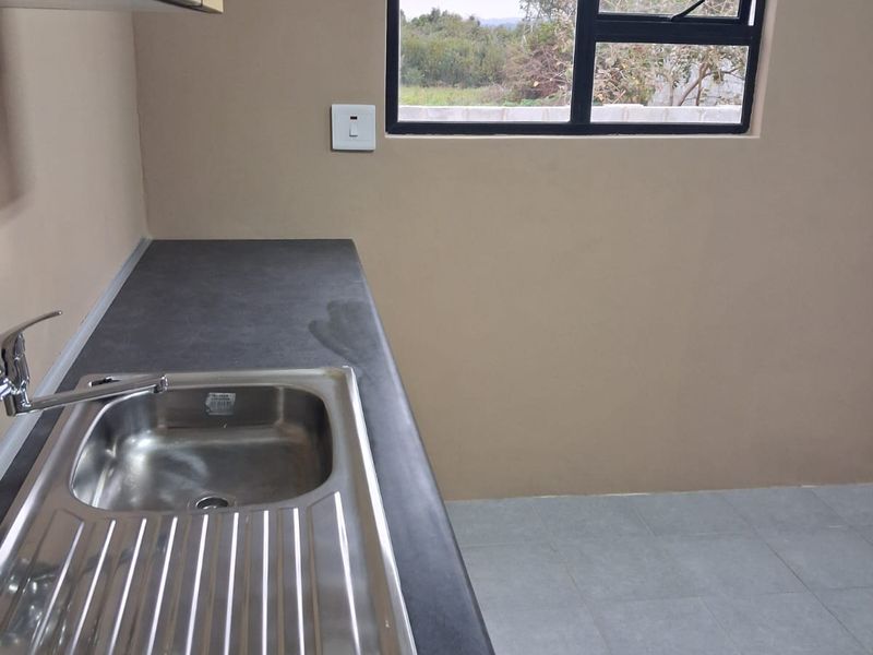A clean, empty kitchen area featuring a stainless steel sink, dark laminate countertop, and tiled flooring with a view through the window.