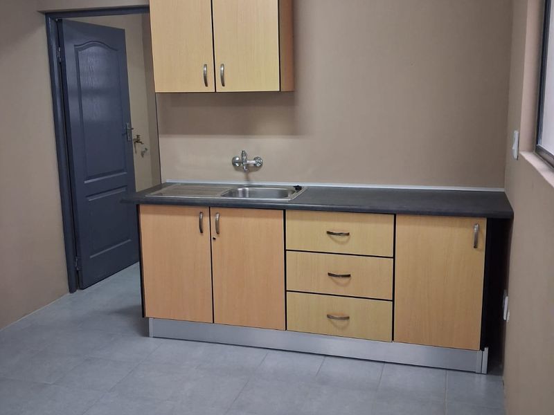 A compact kitchen area featuring light wood-finish cabinetry, a stainless steel sink, dark laminate countertops, and neutral tiled flooring.