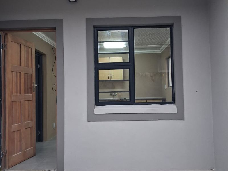 A covered patio area featuring a timber entrance door, aluminium-framed window, and exterior lighting, with an unfinished concrete floor.