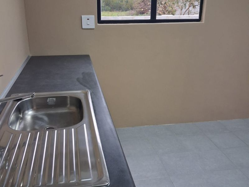 A clean, empty kitchen area featuring a stainless steel sink, laminate countertop, and neutral tiled flooring with a window providing natural light.