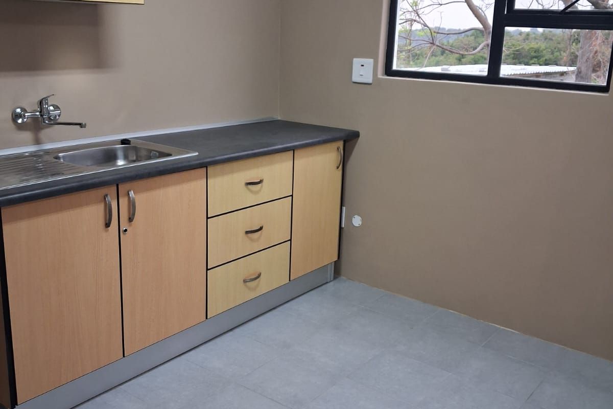 A clean, functional kitchen area featuring light wood-tone cabinetry, dark laminate countertops, a stainless steel sink, and neutral tiled flooring.