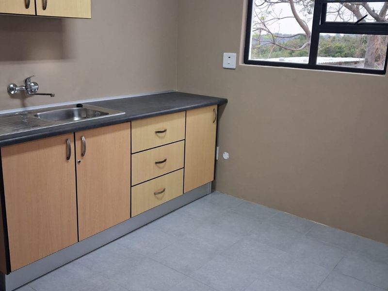 A clean, functional kitchen area featuring light wood-tone cabinetry, dark laminate countertops, a stainless steel sink, and neutral tiled flooring.