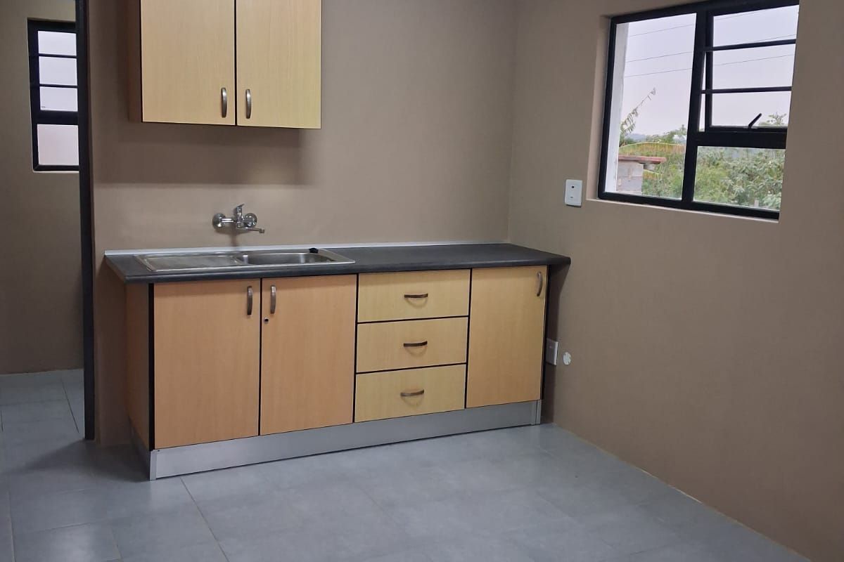 A clean, empty kitchen featuring light wood-tone cabinetry, a stainless steel sink, tiled flooring, and neutral wall finishes.