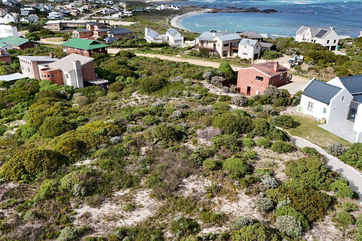 An aerial view of a vacant residential plot featuring natural coastal vegetation and sandy terrain, situated in a developed coastal neighborhood.