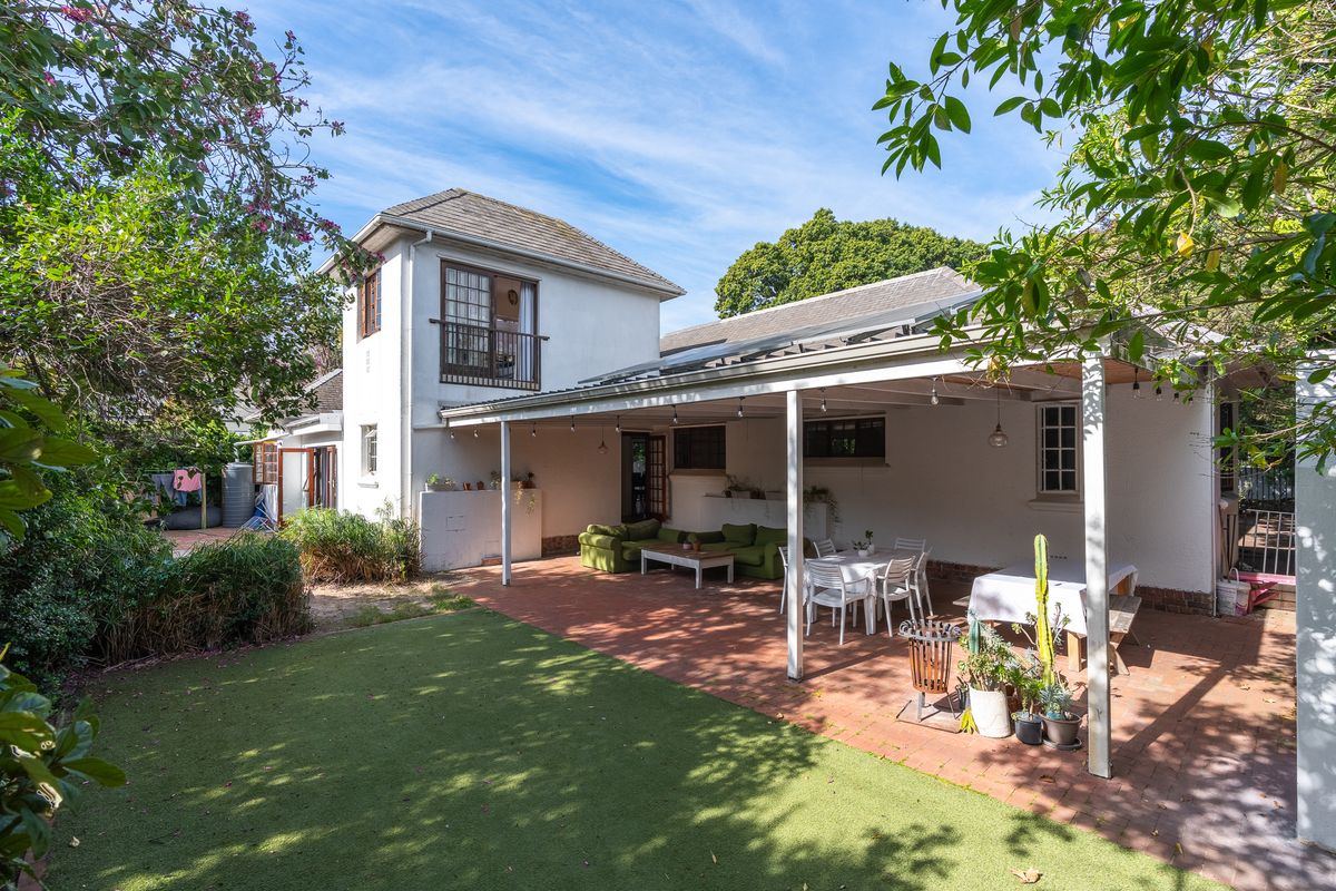 Rear view of the property featuring a spacious covered patio, brick paved area, artificial lawn, and an upper-level balcony.