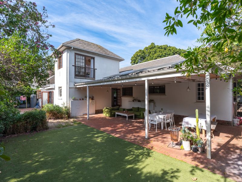 Rear view of the property featuring a spacious covered patio, brick paved area, artificial lawn, and an upper-level balcony.