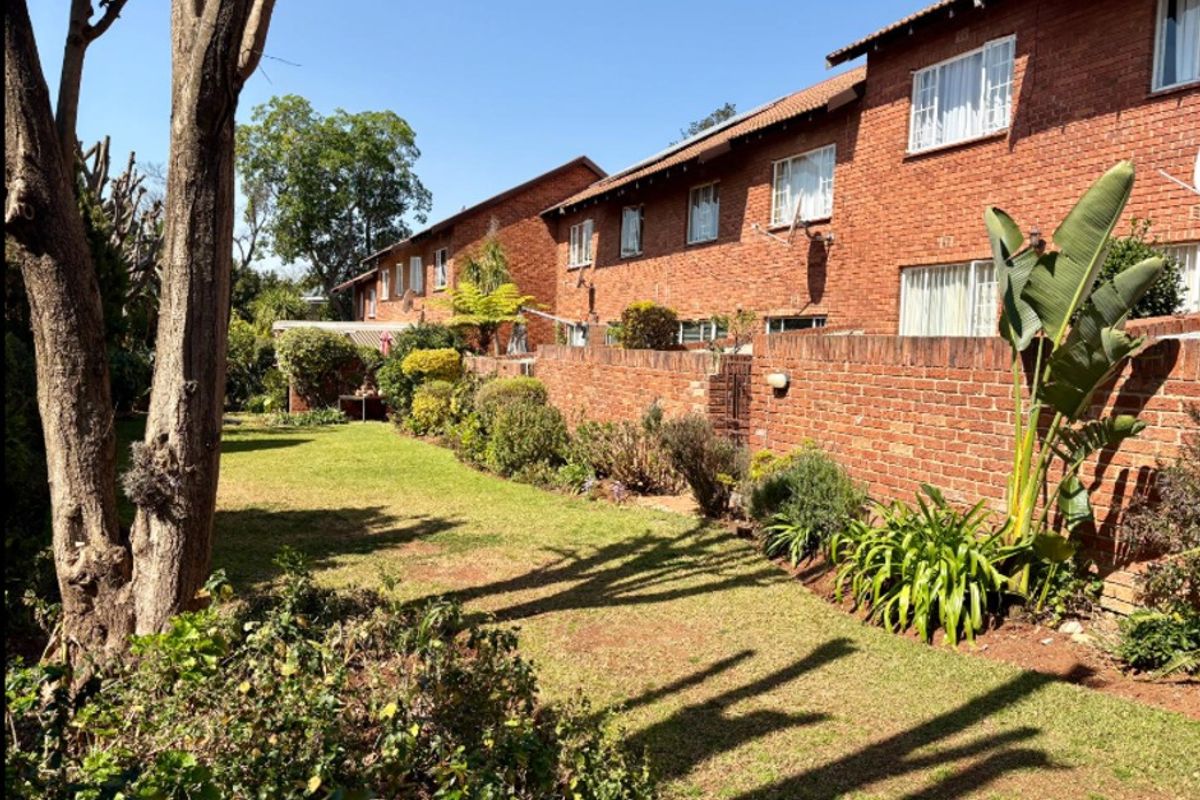 A view of the rear exterior of the townhouse complex, featuring a well-maintained lawn, landscaped garden beds, and a classic face brick facade.