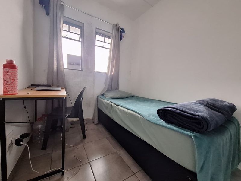 A compact bedroom featuring tiled flooring and dual casement windows, showing signs of wear on the wall surfaces.