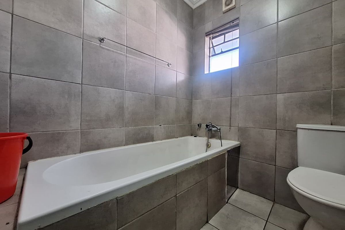 A functional bathroom featuring a built-in bathtub, toilet, and neutral grey wall and floor tiling with an integrated extractor fan.