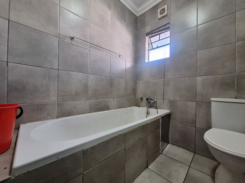 A functional bathroom featuring a built-in bathtub, toilet, and neutral grey wall and floor tiling with an integrated extractor fan.