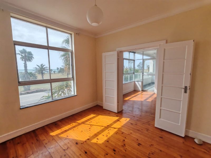 Bright and spacious living area featuring original timber flooring, large windows with coastal views, and double doors leading to an enclosed balcony.