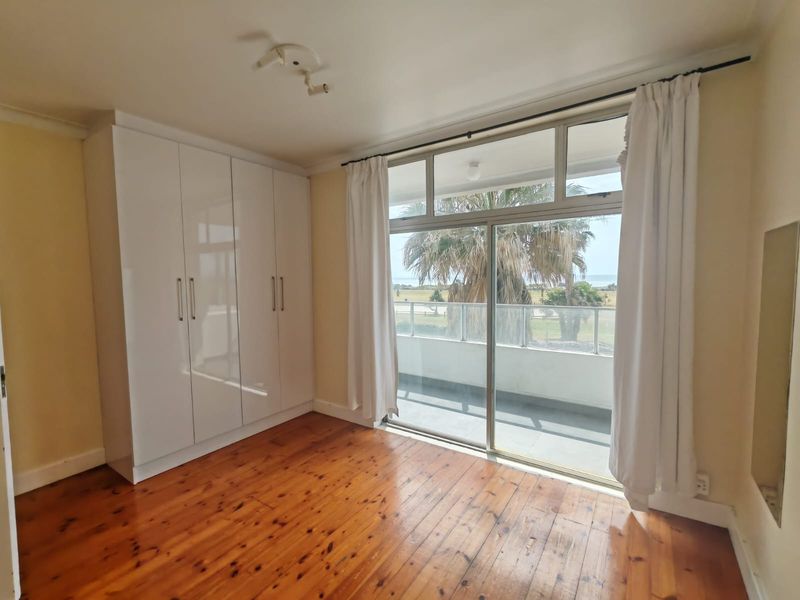 A bright bedroom featuring polished timber flooring, a modern white built-in wardrobe, and sliding glass doors leading to a balcony with coastal views.