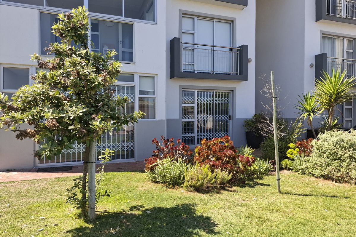 Rear view of the apartment building featuring a landscaped garden, paved walkway, and ground-floor access with security gates and an upper-level balcony.