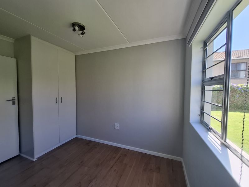 A bright, empty bedroom featuring laminate flooring, built-in cupboards, and large windows with security bars overlooking the garden.
