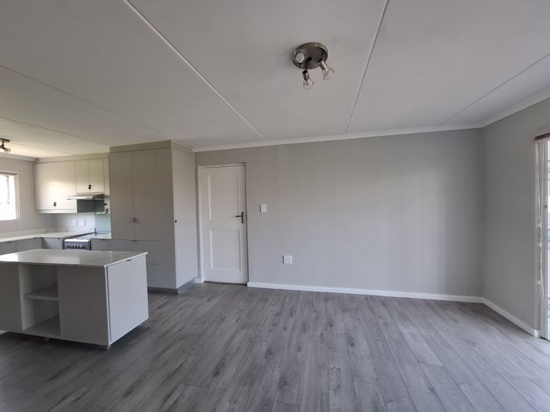A bright open-plan living and kitchen area featuring modern grey laminate flooring, neutral walls, and a functional kitchen with built-in cabinetry and an island.