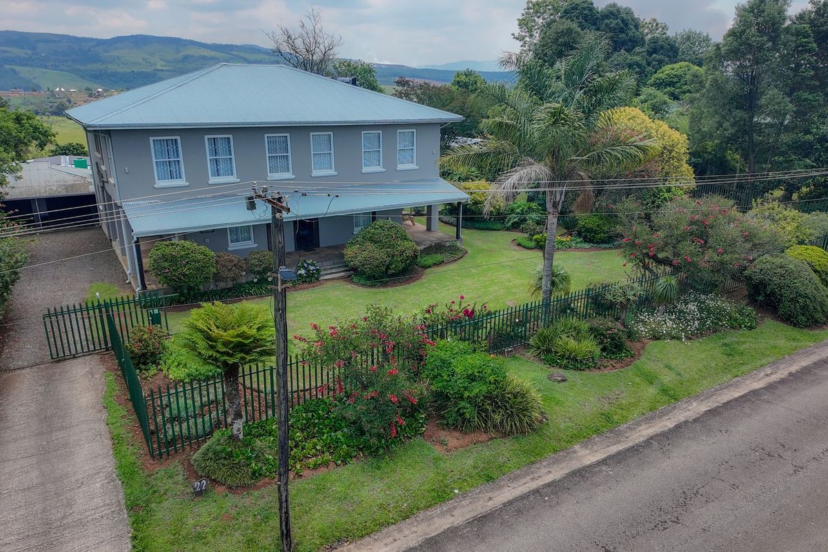 Aerial view of a two-story residential property featuring a metal roof, wrap-around veranda, and a well-maintained landscaped garden with perimeter fencing.