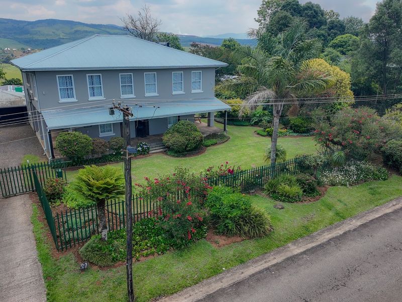 Aerial view of a two-story residential property featuring a metal roof, wrap-around veranda, and a well-maintained landscaped garden with perimeter fencing.